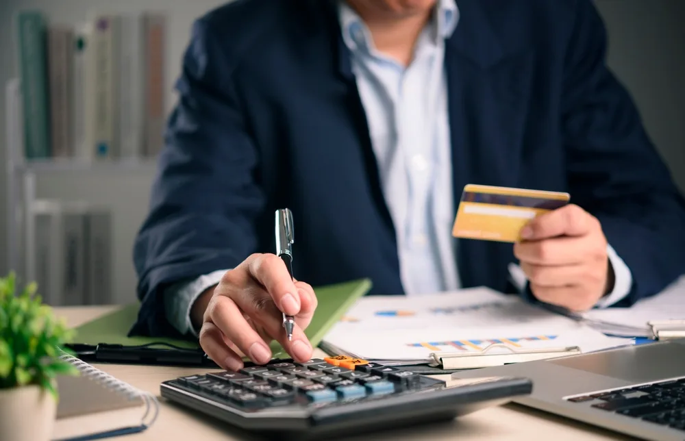 A person seated at a desk, using a calculator while holding a bank card, with paperwork and a laptop in front of them.